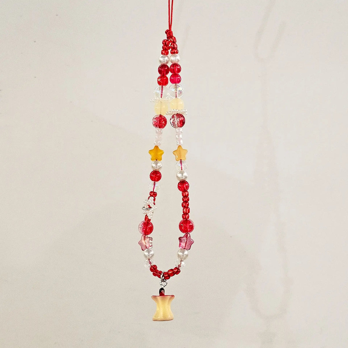 A red beaded hanging ornament with translucent red and clear beads, yellow star-shaped beads, and a small yellow bell-shaped charm at the bottom, shown against a plain off-white background.