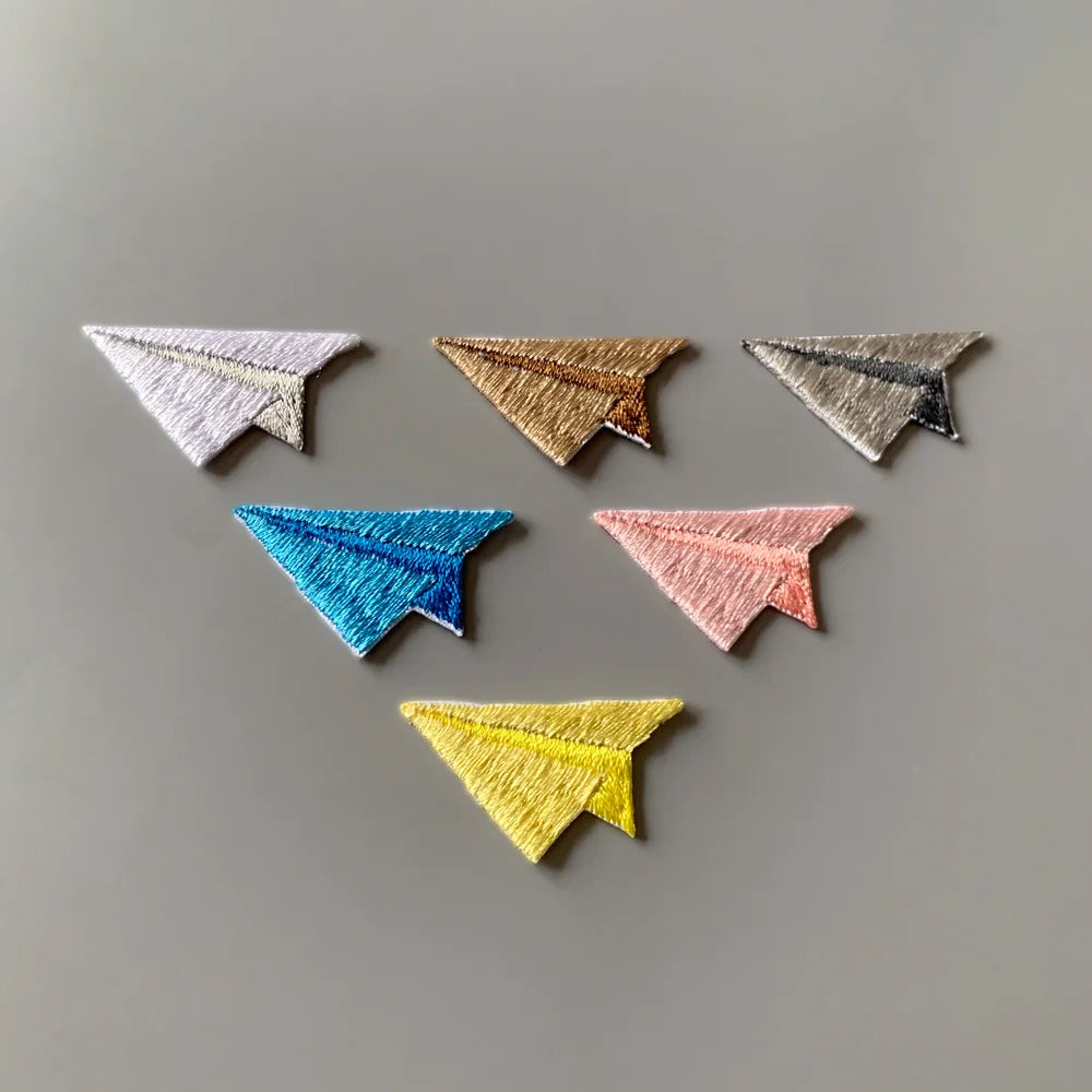 Six embroidered paper airplane patches in different colors (white, gold, silver, blue, pink, yellow) arranged on a neutral gray background.