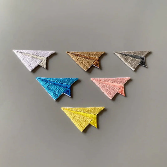 Six embroidered paper airplane patches in different colors (white, gold, silver, blue, pink, yellow) arranged on a neutral gray background.