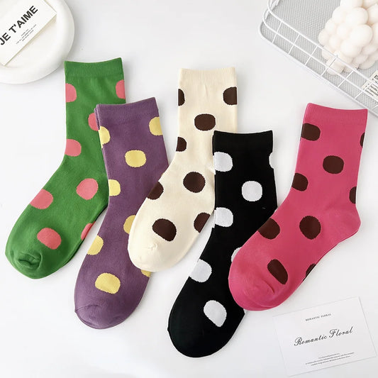 Five pairs of ankle socks arranged in a flat lay on a white surface, each with large polka dots in different colors: green with pink and yellow dots, purple with yellow dots, cream with brown dots, black with white and brown dots, and pink with brown dots.