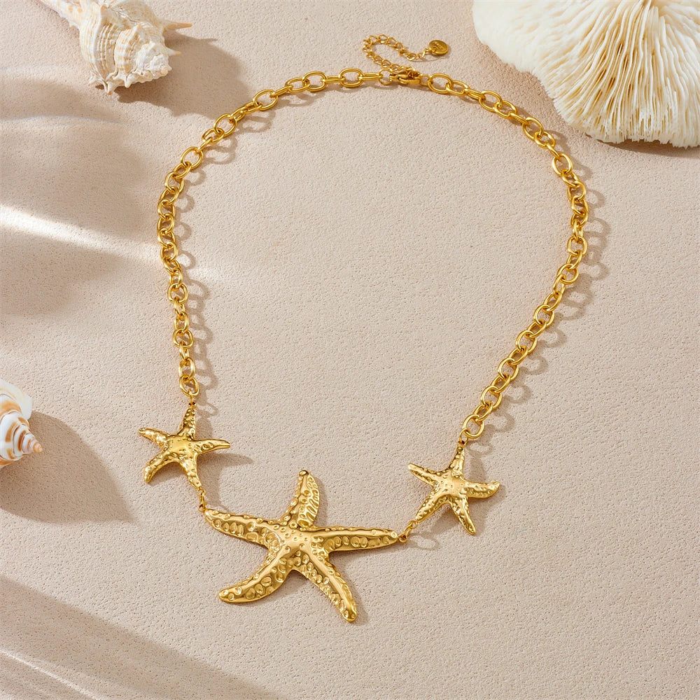 A gold-toned chain necklace featuring three textured starfish charms, displayed on a beige surface with decorative seashells.