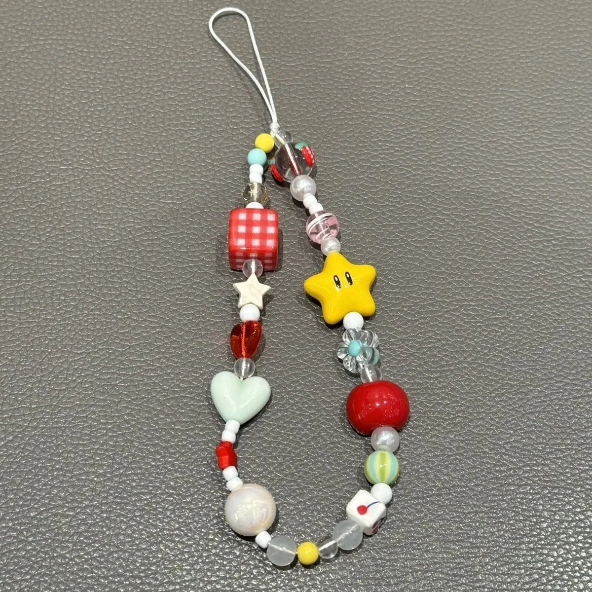 A colorful beaded charm strap with a white plastic loop, featuring various cartoon and gaming-inspired beads including a yellow star, a red gingham cube, a mint green heart, and red and yellow accents, displayed on a textured gray background.