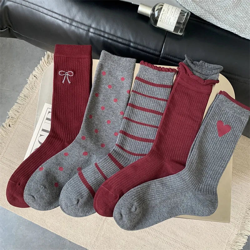 Four pairs of ribbed crew socks arranged in a fan shape on a light beige surface. The set includes gray socks with red hearts, red bows, red stripes, and a red heart, alongside solid dark red pairs.