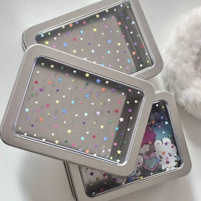 Three square metal tin boxes with clear lids decorated with colorful star stickers, stacked on a light surface with a fluffy white prop on the right.