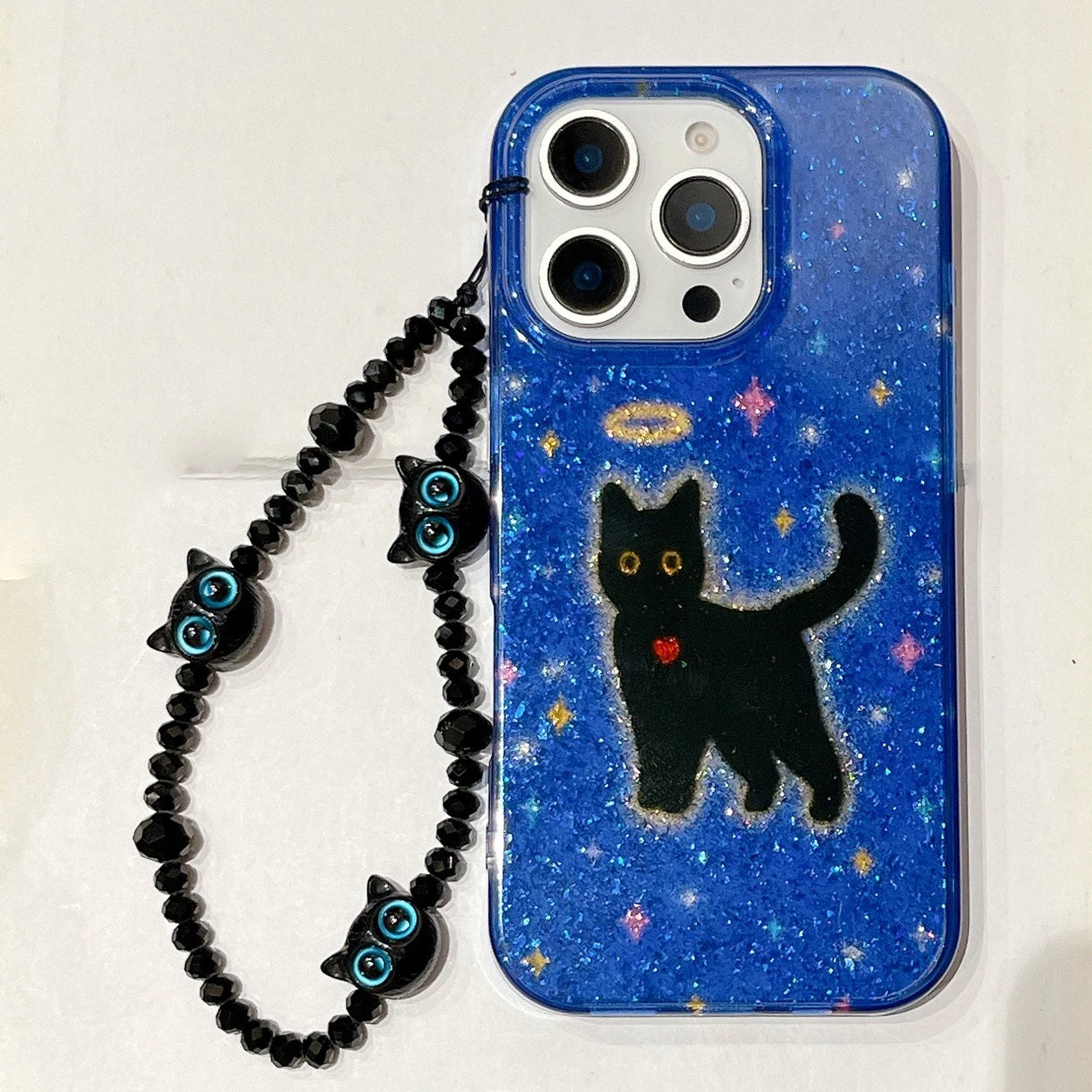A blue glittery phone case with a black cat illustration, gold halo, and small red heart, attached to a black beaded wrist strap with blue eye charms, shown on a white background.