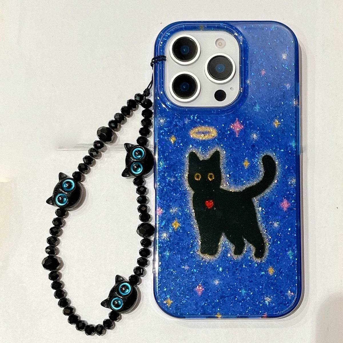 A blue glittery phone case with a black cat illustration, gold halo, and small red heart, attached to a black beaded wrist strap with blue eye charms, shown on a white background.