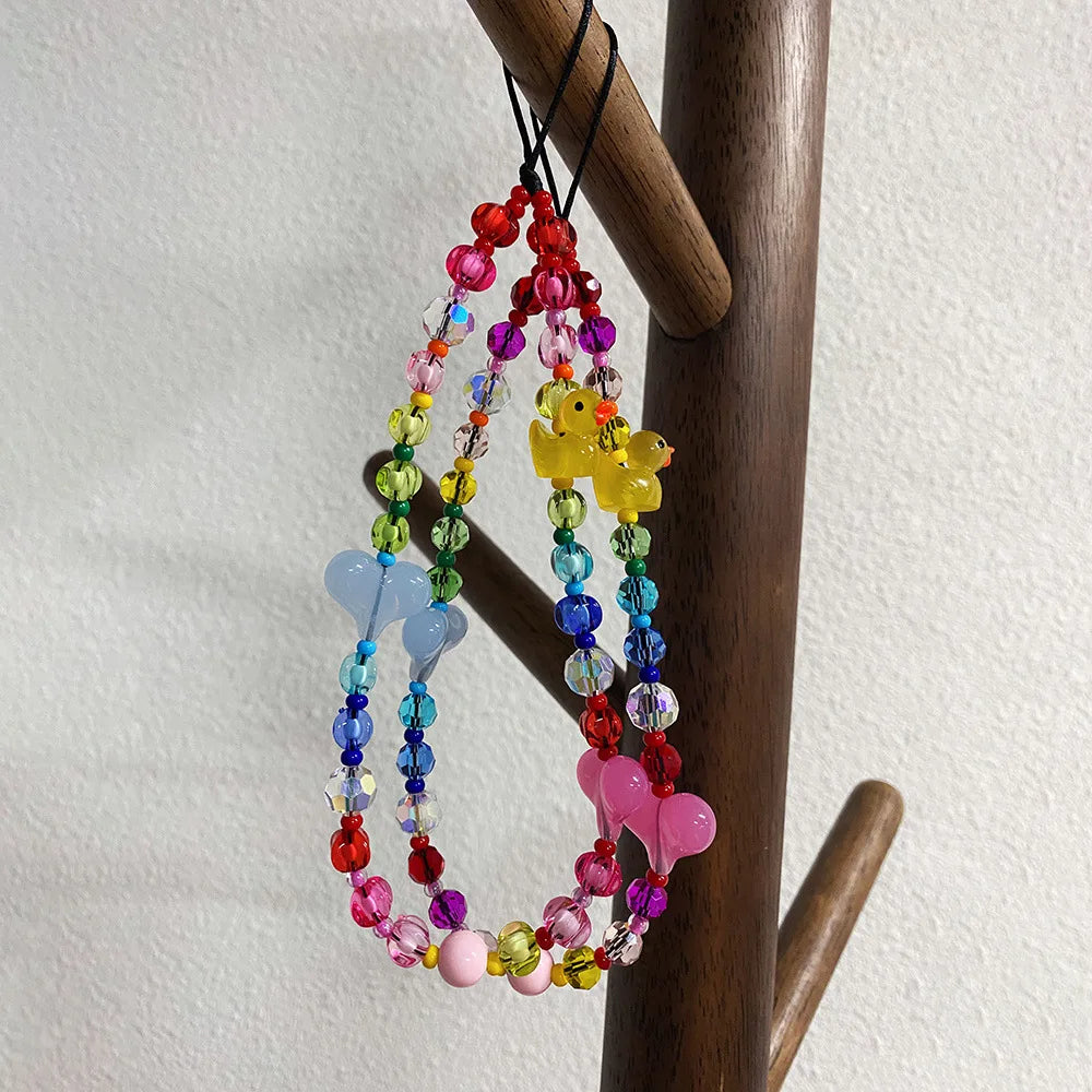 A colorful beaded phone strap or necklace accessory hanging from a wooden coat rack. The accessory features multicolored faceted beads, a translucent light blue butterfly charm, a yellow duck charm, and a pink butterfly charm.