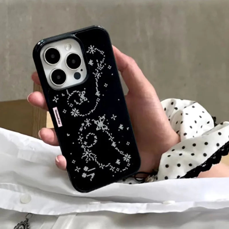 A hand holding a black phone case decorated with a white constellation pattern, set against a background of a white polka-dot shirt and a neutral wall.