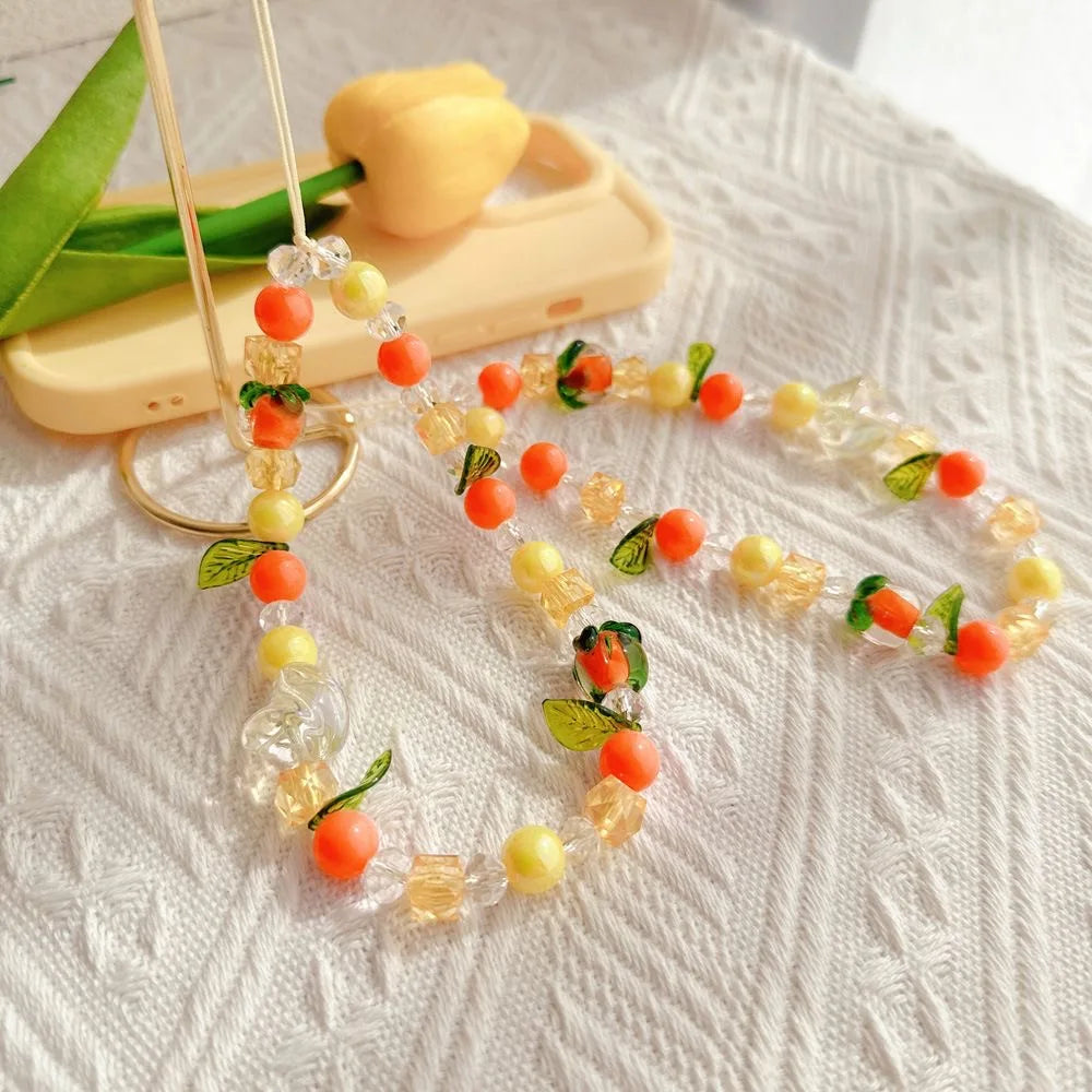 A beaded phone strap or necklace with clear faceted beads, orange pom-pom accents, yellow beads, and small green leaf details, displayed on a textured white fabric with a cream phone case and tulips in the background.