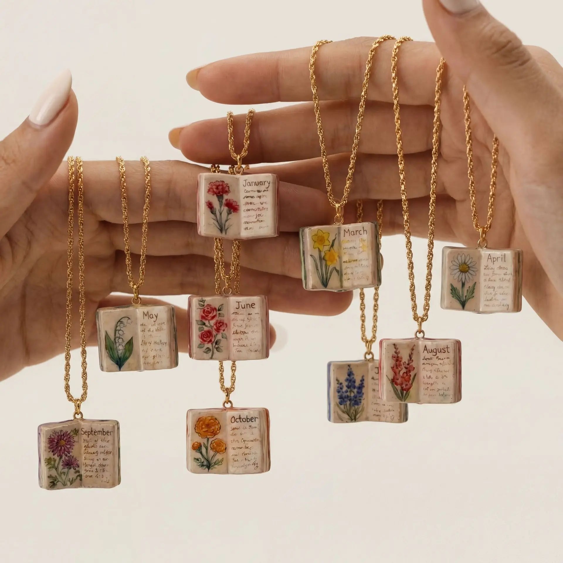 Hand holding necklaces with small book pendants featuring floral designs and months of the year text.