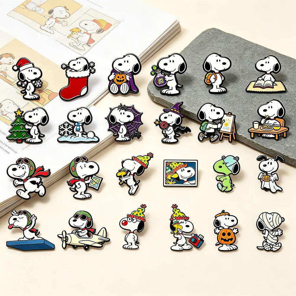A set of Snoopy enamel lapel pins arranged on a light beige surface with styled props, including an open book and a gray stone slab. The pins feature multiple cartoon designs of Snoopy in various activities and costumes, including Christmas, Halloween, and everyday scenes.