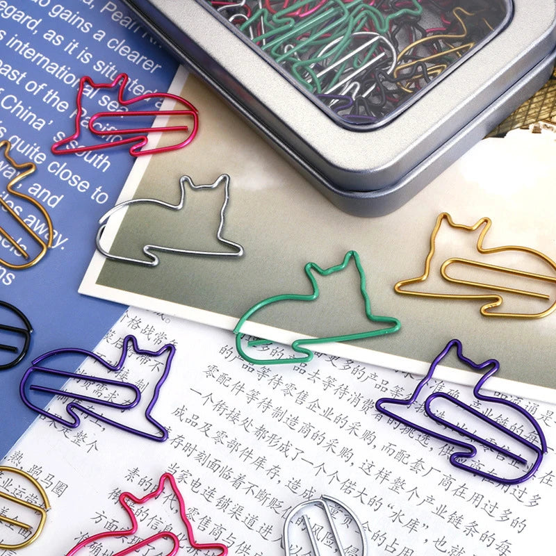 A set of colorful cat-shaped wire paperclips in silver, gold, green, blue, red, and purple, displayed on top of printed papers with a metal tin of paperclips in the background.