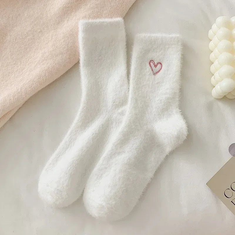 A pair of fluffy white socks with a small pink embroidered heart on the cuff, styled on a light surface with soft neutral props.