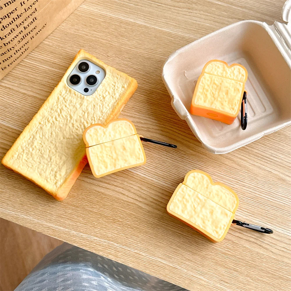 A set of bread-topped phone case and wireless earbuds cases in a warm yellow and orange gradient, styled on a light wood table with a paper coffee cup and a phone visible.