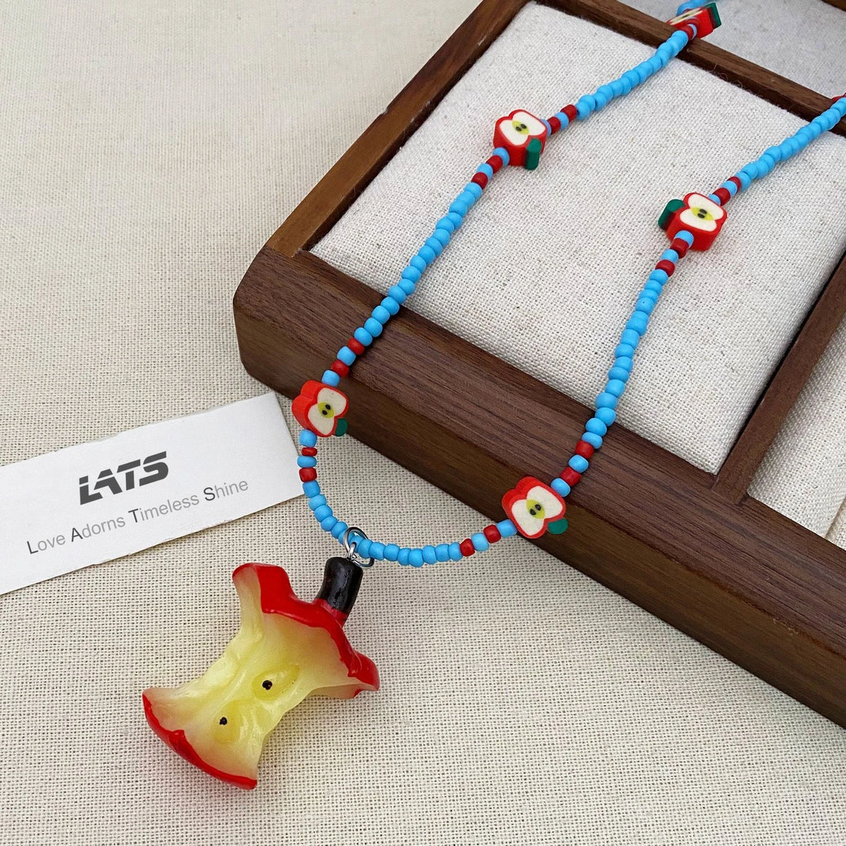 A beaded necklace with bright blue beads, small red and white flower accents, and a translucent red-and-yellow apple-bite pendant, displayed on a cream fabric surface with a wooden tray and a card reading LATS.