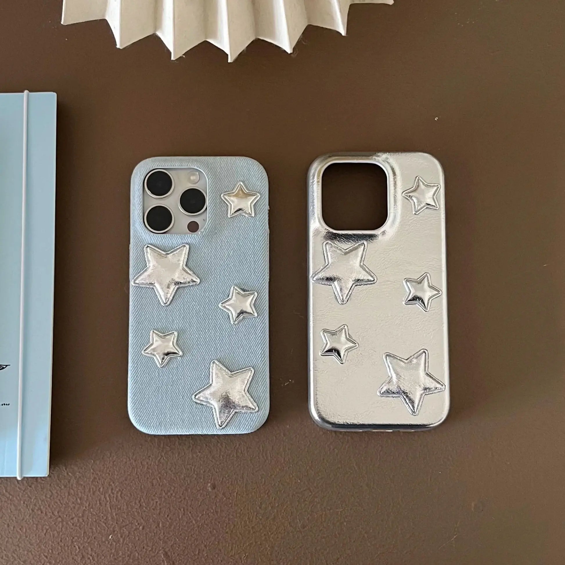 Two phone cases with raised star designs, displayed side by side on a brown surface. The left case is light blue with silver stars, and the right case is metallic silver with silver stars.