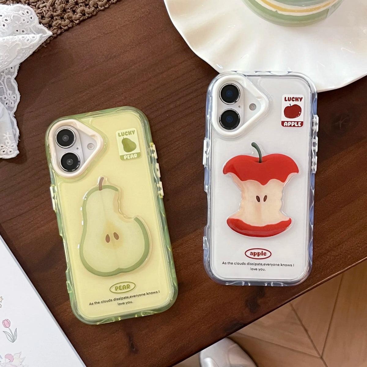 Two clear phone cases on a wooden table. One case is pale yellow with a 3D pear design and 'LUCKY PEAR' text.