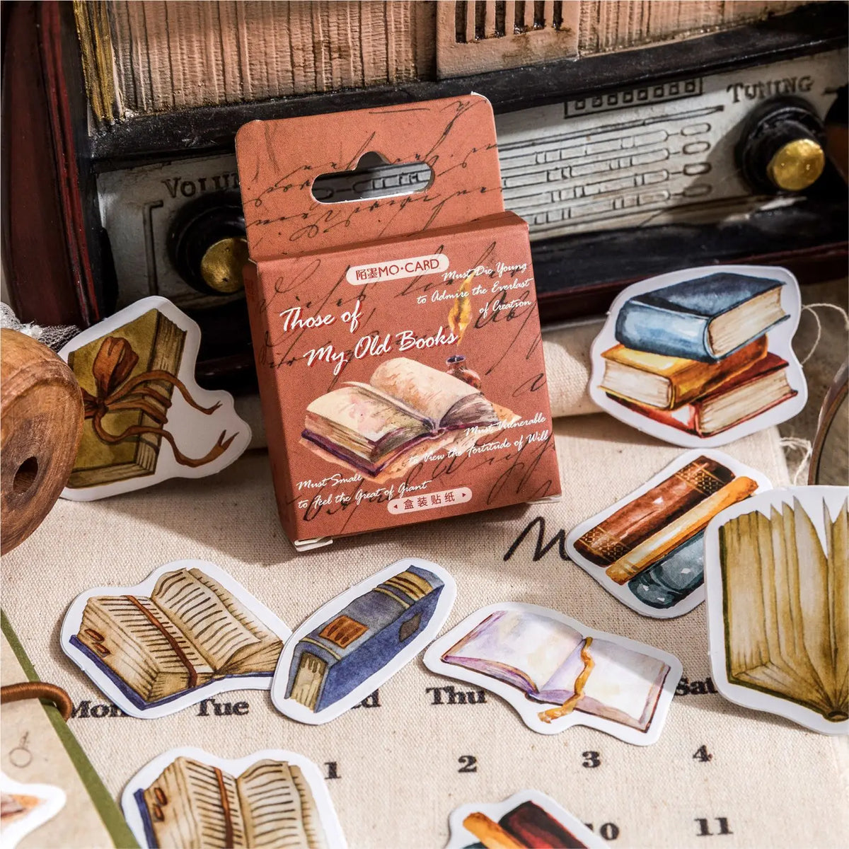 A vintage-style card set box labeled 'NEMO CARD Those of My Old Books' stands on a fabric surface, surrounded by illustrated book sticker/card cutouts and an antique radio in the background.