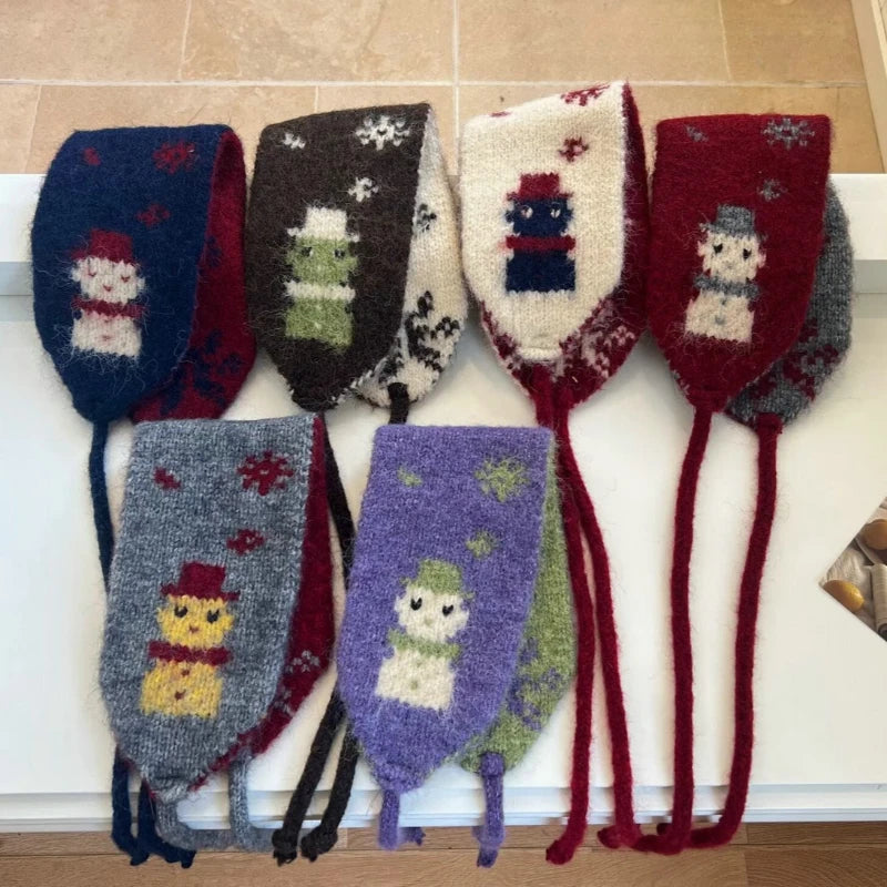 Six fuzzy scarves with long ties, arranged on a white surface. Each pair has a different winter-themed knit design, including snowmen, snow globes, and Christmas trees in colors like navy, brown, cream, red, gray, purple, and green.