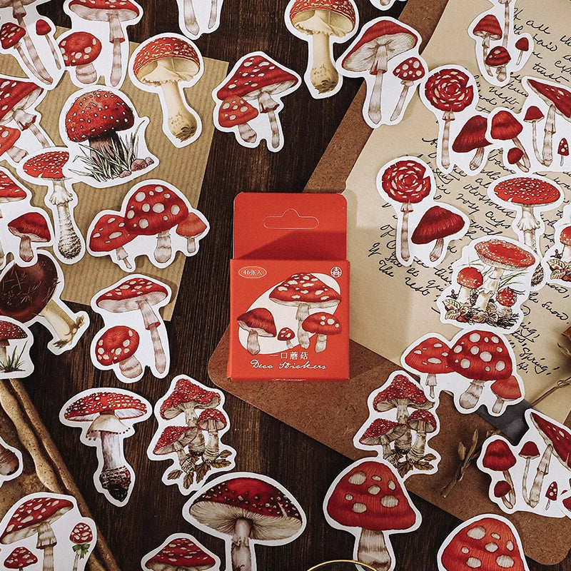 A styled flat-lay of red and white mushroom stickers scattered across a dark wood surface, with a small red box labeled 'Pure Stickers' in the center. Decorative props include vintage-style papers with cursive writing, a wooden spoon, and a cinnamon stick.