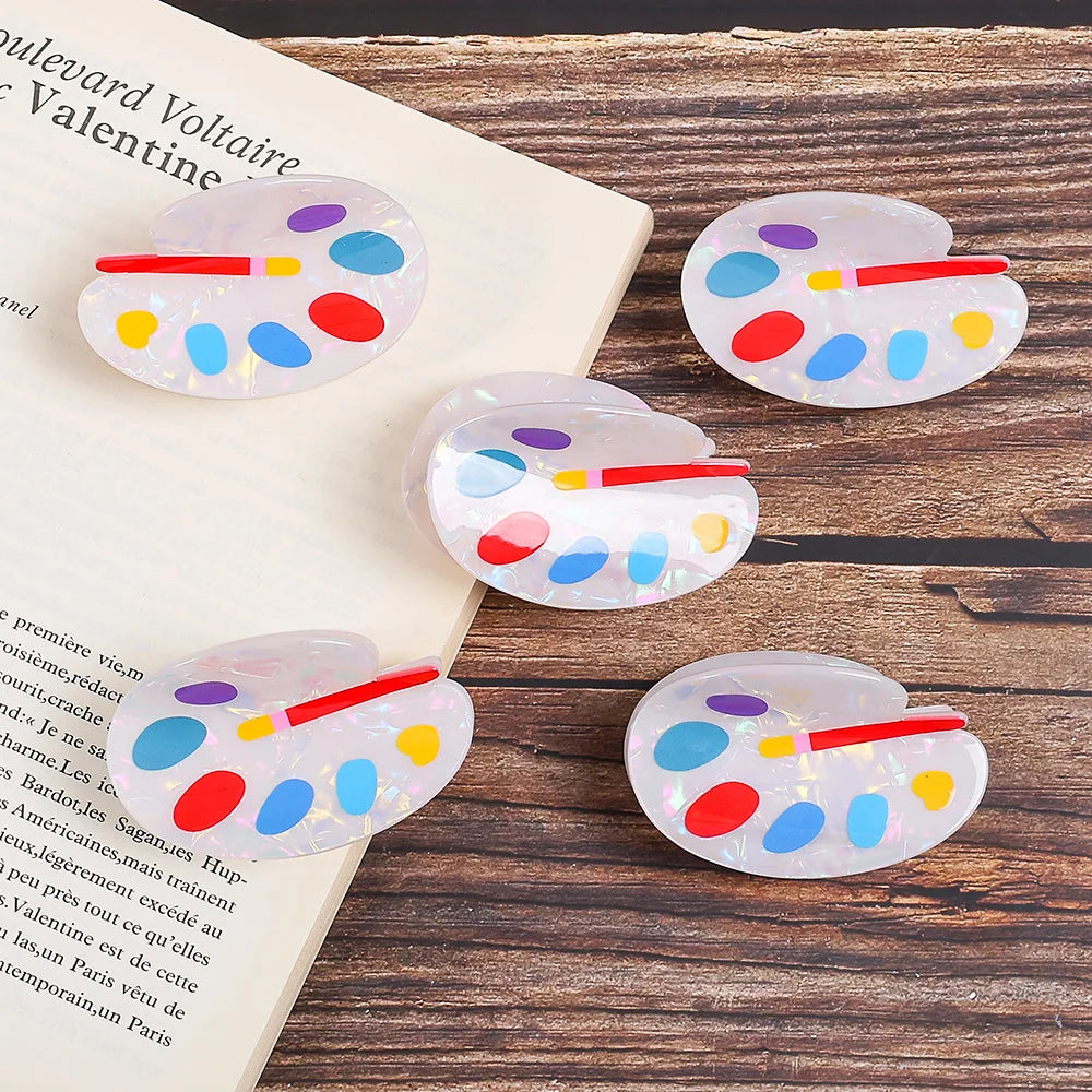 Five acrylic hair claw clips designed like miniature paint palettes, displayed on a wooden surface next to an open book. The clips are made of translucent white acrylic with colorful painted dots in red, blue, yellow, and purple, and a small red handle.