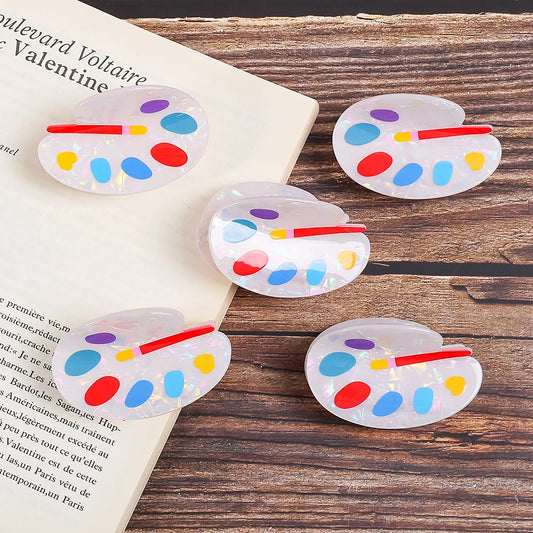 Five acrylic hair claw clips designed like miniature paint palettes, displayed on a wooden surface next to an open book. The clips are made of translucent white acrylic with colorful painted dots in red, blue, yellow, and purple, and a small red handle.