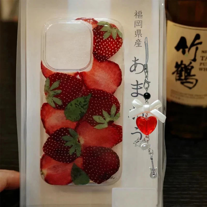 A clear phone case printed with a strawberry slice pattern, shown inside its white retail packaging. The package includes a decorative charm with a red heart, white bow, and silver guitar, alongside Japanese text.
