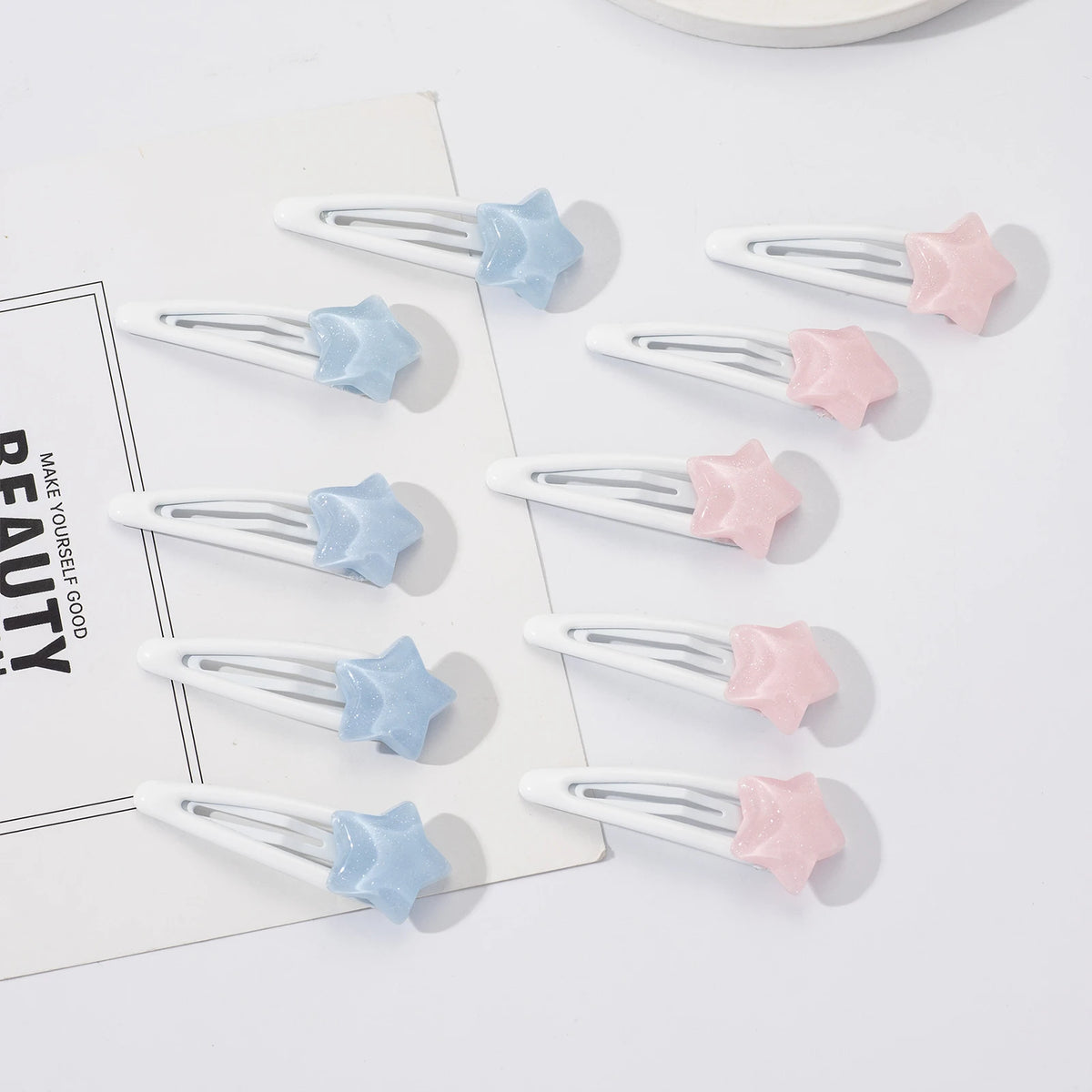 Nine white hair claw clips with star-shaped ends in pastel blue and pink, arranged on a white surface with a decorative card.