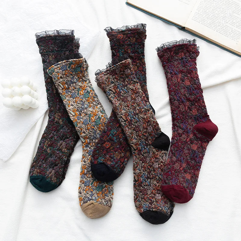 Four pairs of floral lace socks arranged on a white fabric surface with a book page and small white decorative object as props. The socks feature multicolored floral patterns in dark, muted tones with black, tan, and burgundy accents.