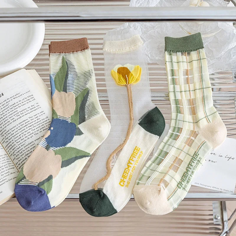 Three pairs of decorative crew socks displayed on a metal shelf with styled props, including an open book and a white plate. The socks feature floral prints, a yellow flower motif, and plaid patterns in soft pastel tones.