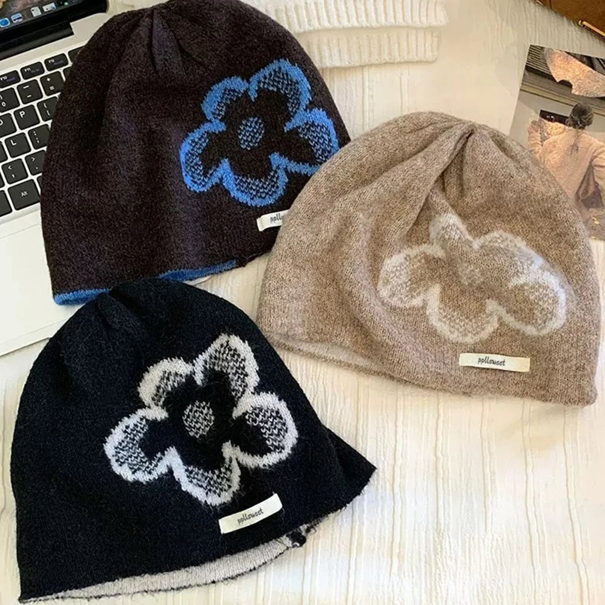 Three knit beanie hats arranged on a light fabric surface: one dark brown with a blue flower motif, one taupe-beige with a white flower motif, and one very dark navy with a white flower motif. Each hat has a small sewn label.