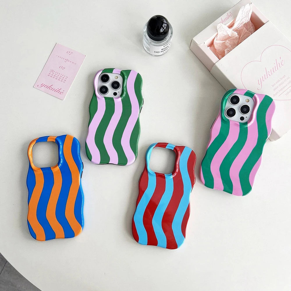 Four wavy-striped phone cases arranged on a white surface, each with a different color combination: blue and orange, green and pink, red and blue, and green and pink. A small pink card, a black-capped bottle, and a white gift box are placed nearby.