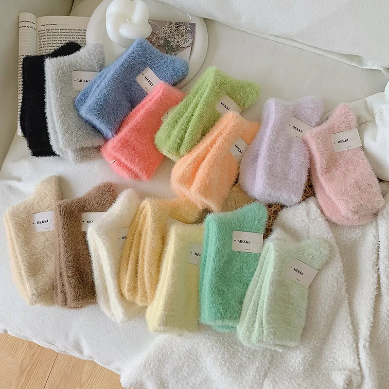 A set of soft, fuzzy socks in multiple pastel and neutral colors, including black, gray, blue, pink, green, orange, purple, beige, brown, cream, and mint, arranged on a white textured blanket with a book in the background.