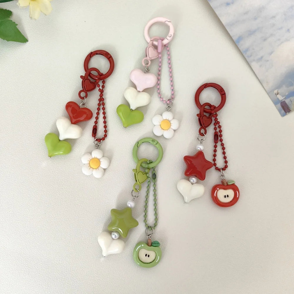 Four colorful acrylic keychain charms with heart, flower, star, and apple shapes, arranged on a light off-white surface.