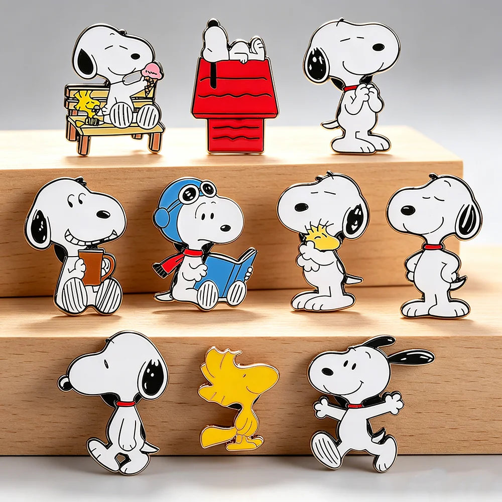 Ten Snoopy enamel pins in various poses are arranged on three wooden display blocks against a light gray background. The pins feature Snoopy and Woodstock in different scenes, including reading, flying a plane, sitting on a bench, and standing.