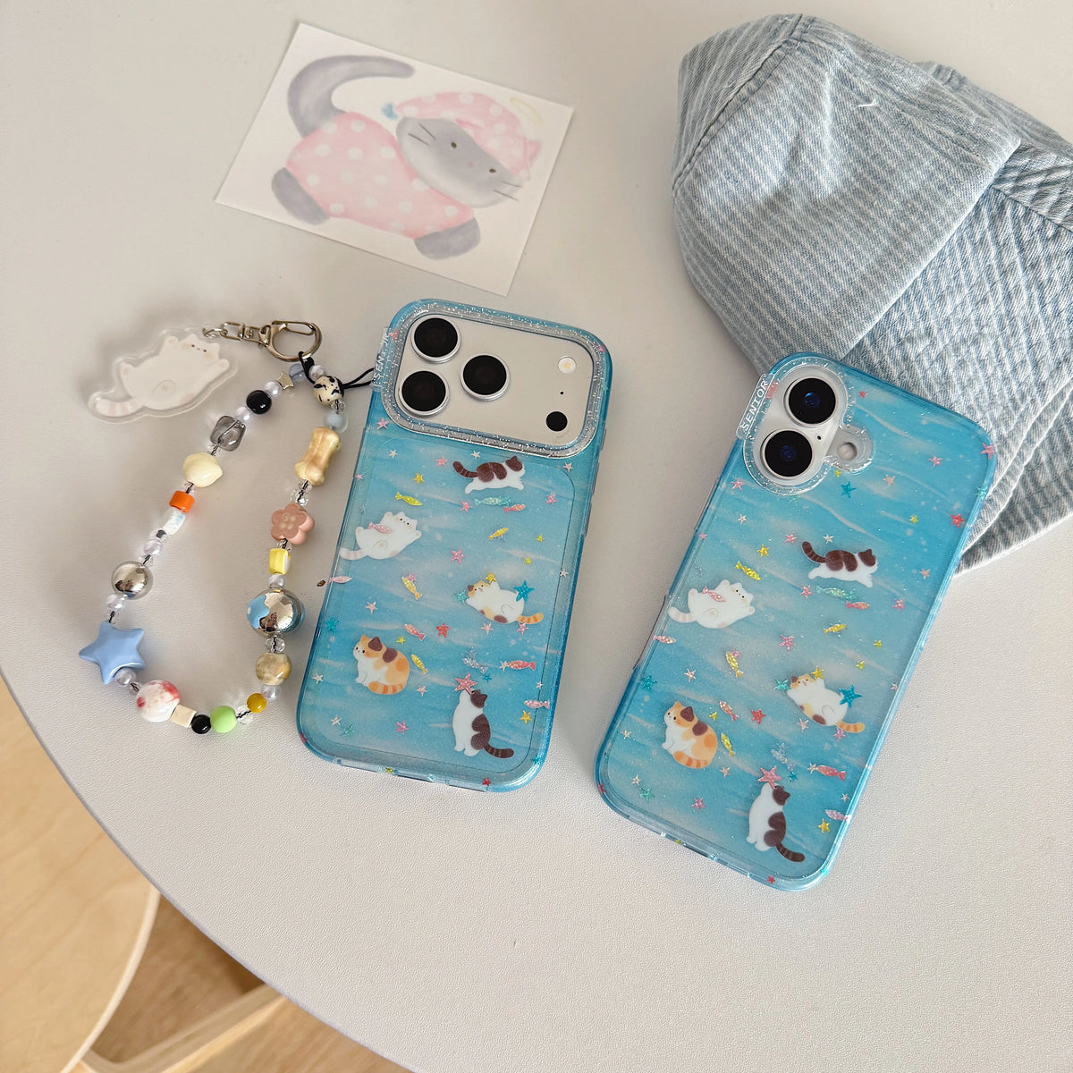 Two light blue phone cases with cute cat illustrations are displayed on a white table alongside a beaded phone strap, a small card, and a striped fabric accessory.