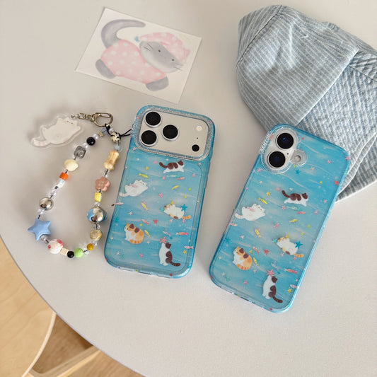 Two light blue phone cases with cute cat illustrations are displayed on a white table alongside a beaded phone strap, a small card, and a striped fabric accessory.