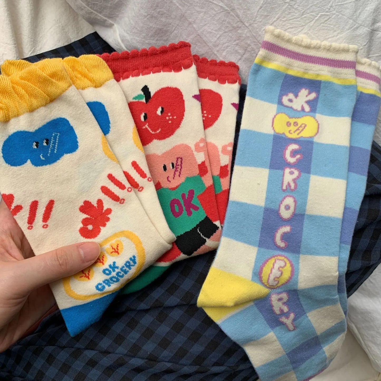 A hand holds a fan of four colorful novelty socks with cartoon graphics and text, laid over a dark plaid fabric background.