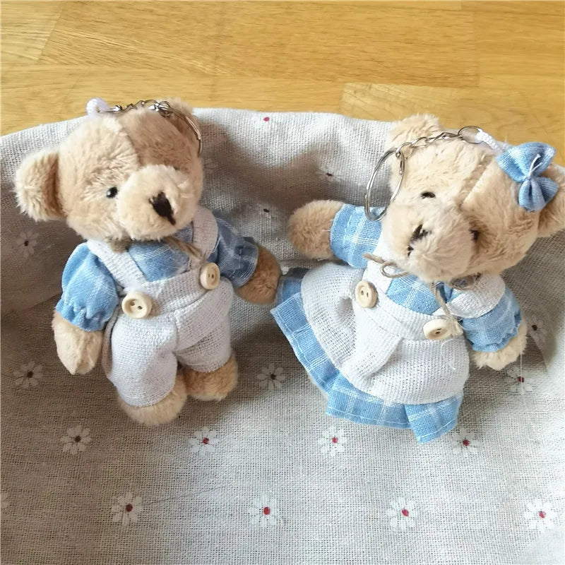Two small plush teddy bear keychains dressed in blue and white outfits, placed on a light fabric surface with a wooden floor in the background.