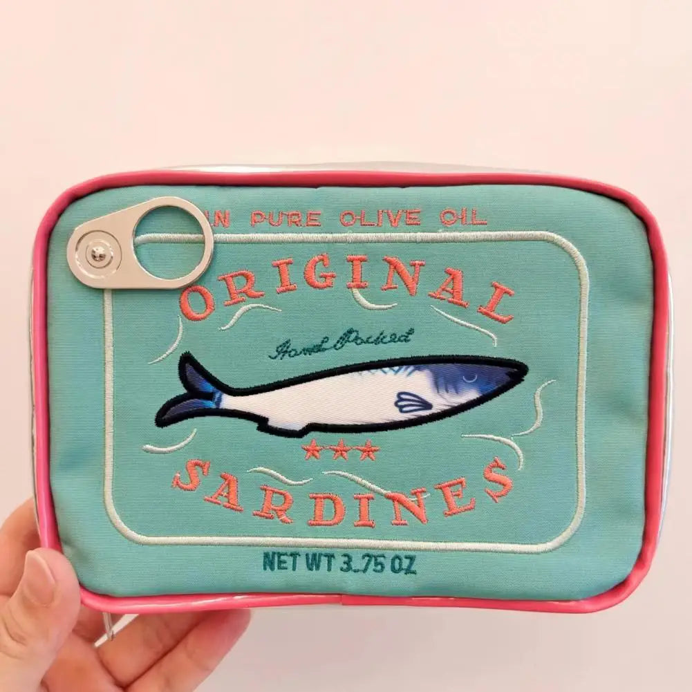A hand holding a small fabric pouch designed to look like a vintage can of 'Original Sardines' olive oil, featuring a turquoise background, pink border, and a blue-and-white sardine illustration.