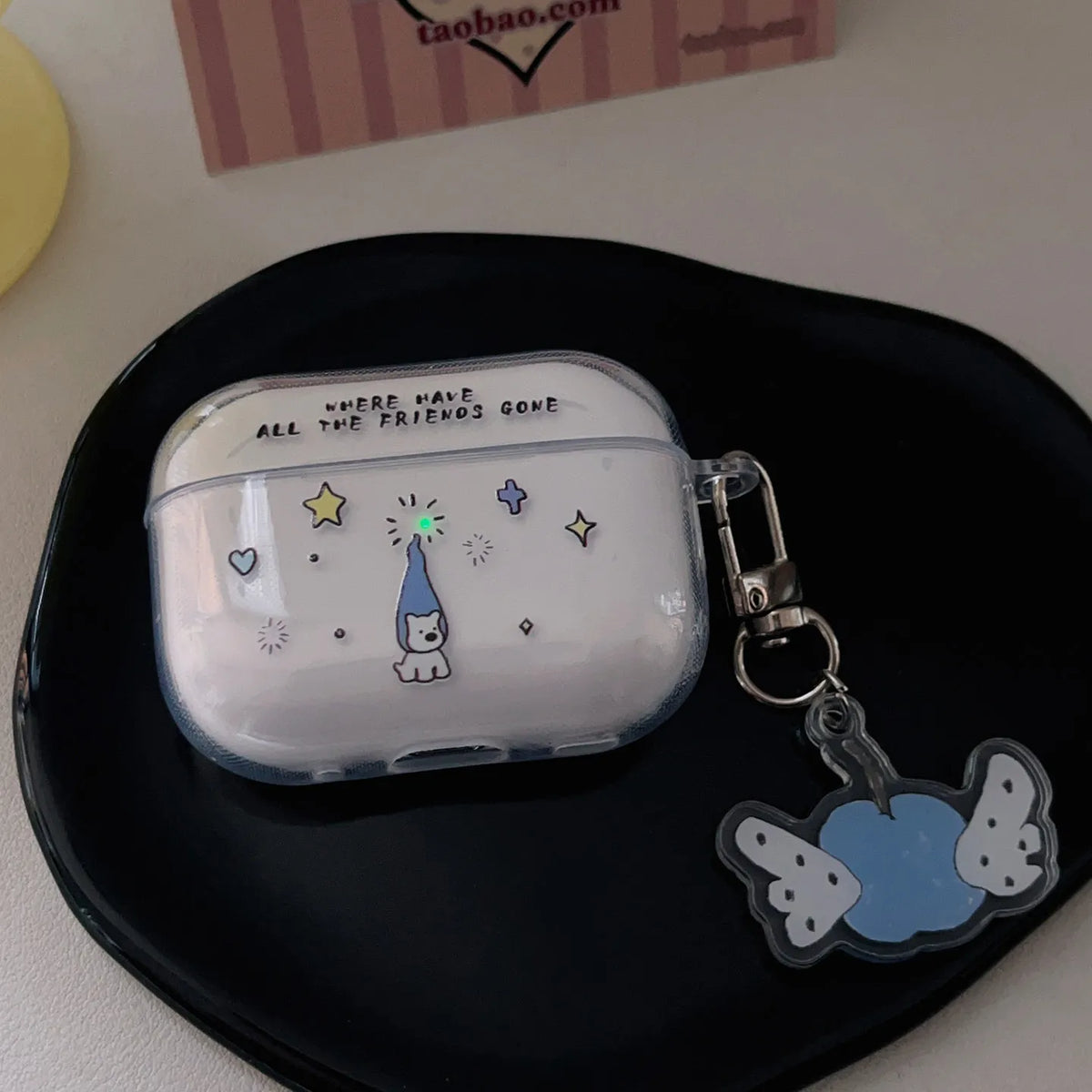 A clear plastic AirPods case with a white interior, decorated with a cute cartoon cat wearing a blue party hat and surrounded by stars and hearts. The case has black text reading 'WHERE WERE ALL THE FRIENDS GONE' on the top.