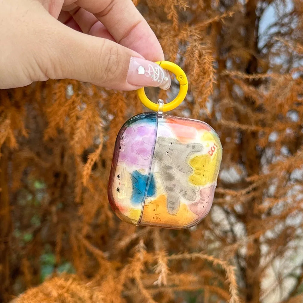 A hand holds a clear AirPods case decorated with colorful pastel cat illustrations and a yellow keychain ring, set against a blurred background of orange-brown foliage.