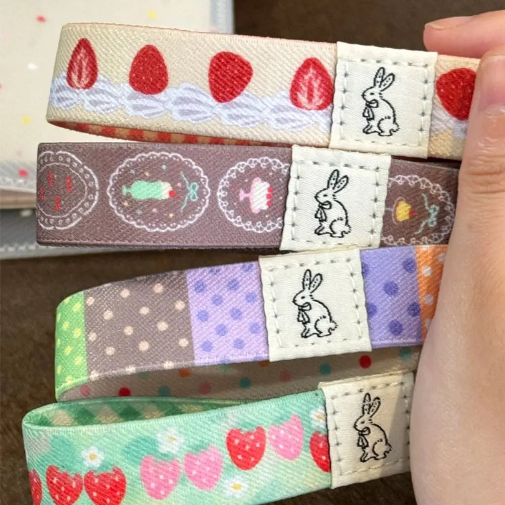 Four colorful patterned notebook strap held in a hand. The book strap feature various pastel and bright patterns including strawberries, cakes, polka dots, and floral motifs, with a white patch on each displaying a black line-art bunny.