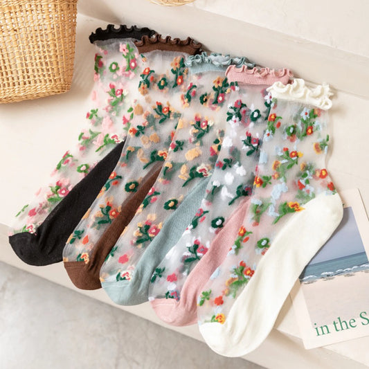 Five pairs of sheer floral embroidered ankle socks with ruffled trim, arranged on a light surface. The socks come in black, brown, mint green, blush pink, and white, each featuring colorful floral embroidery.