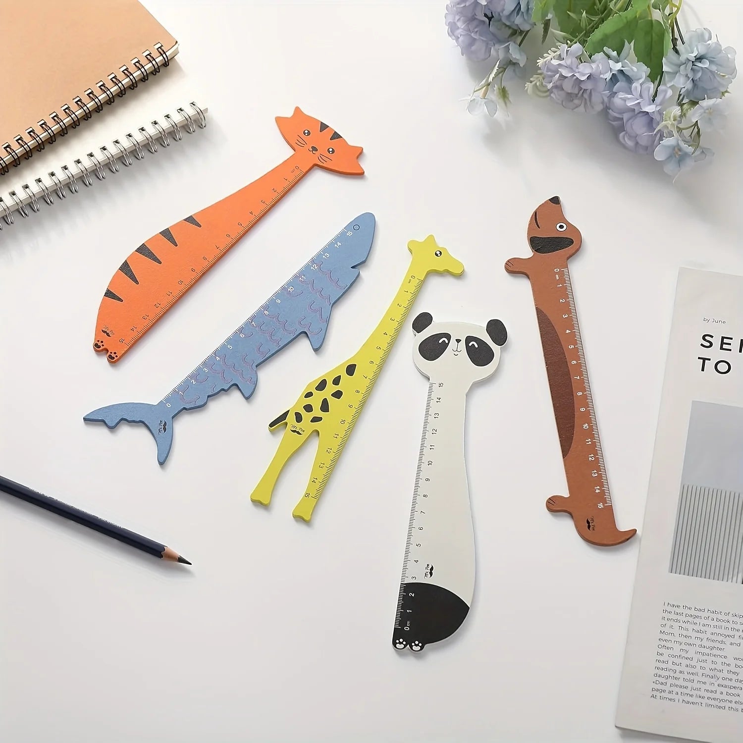 Five animal-shaped rulers on a white surface with a pencil and flowers.