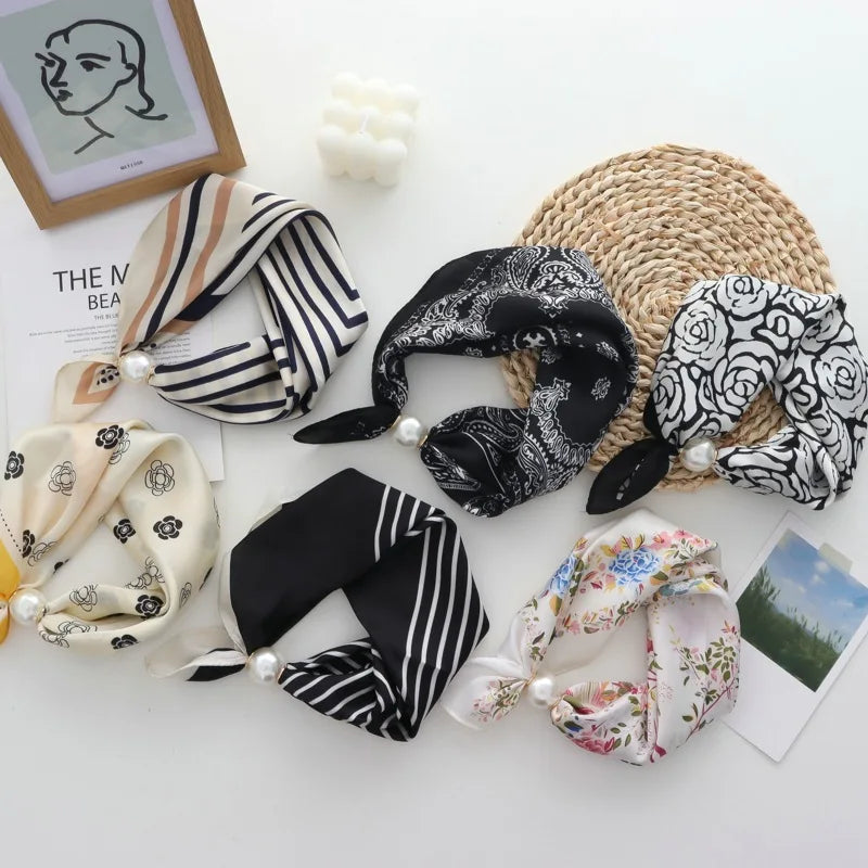 Six different silk scarf headbands with pearl accents are arranged in a flat-lay on a white surface. The designs include stripes, florals, and paisley in black, white, beige, and pastel colors.