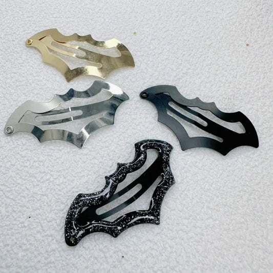 Four bat-shaped metal hair claw clips in different finishes (gold, silver, matte black, and glittery black) arranged on a textured white background.
