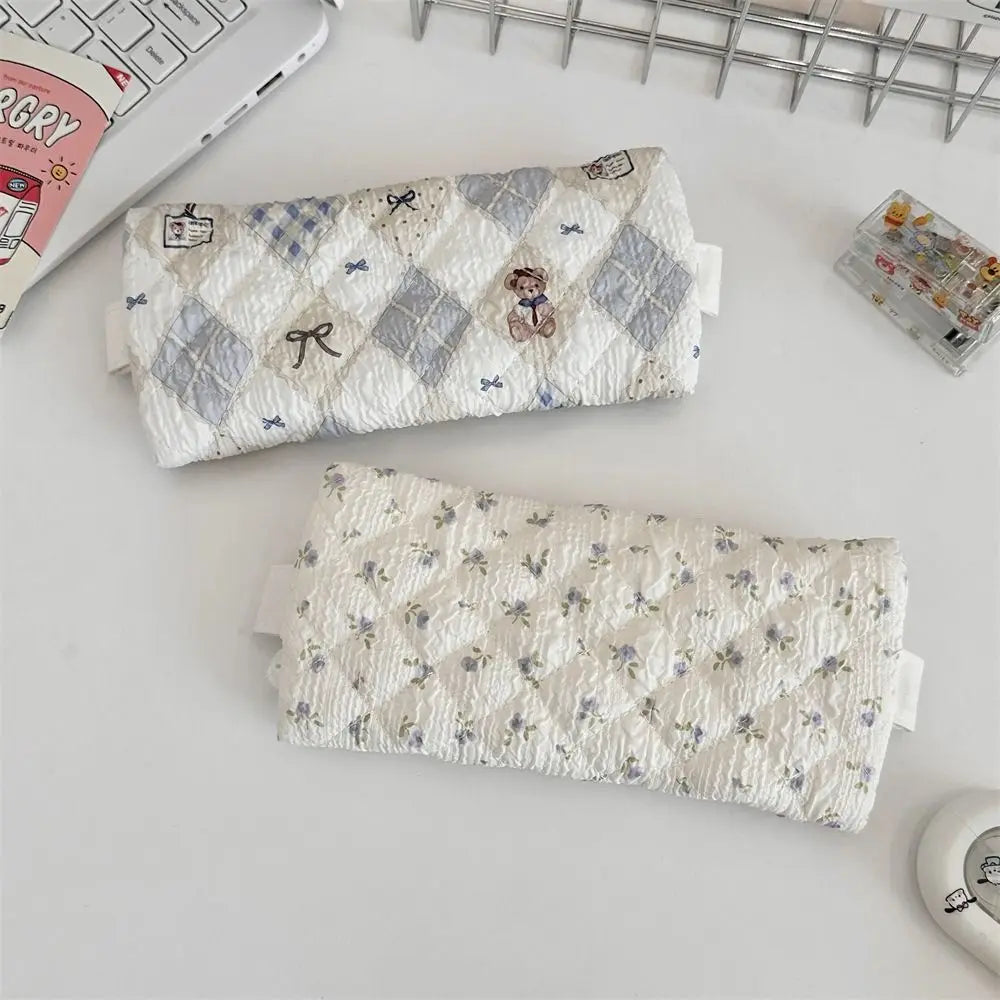 Two quilted pencil cases with textured fabric, one featuring a gray and white argyle pattern with small bows and a teddy bear, and the other with a white background and small blue floral print, displayed on a desk next to a keyboard and stationery.