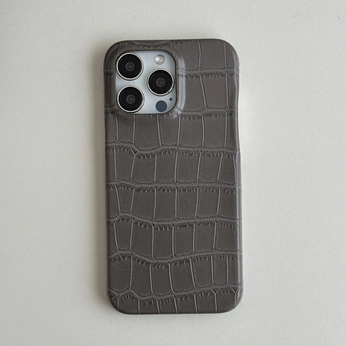 A dark gray phone case with an embossed crocodile skin texture, shown on a silver iPhone against a light gray background.