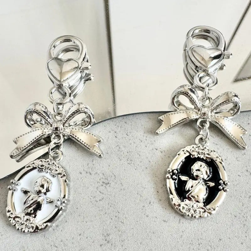 A pair of silver-toned clip-on charms featuring a bow and oval cherub pendant charms, displayed on a textured gray surface.